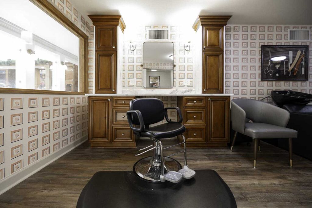 A modern barbershop interior with a single black barber chair in front of a mirror, wooden cabinets, patterned wallpaper, a gray lounge chair, and a hair washing station. The floor is wooden and the space is well-lit.