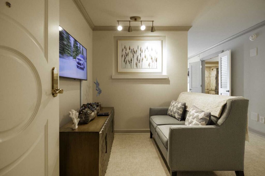 A cozy living room with a gray sofa, patterned pillows, a wooden sideboard, wall-mounted TV, framed artwork, and soft lighting on beige walls and carpeted floor.