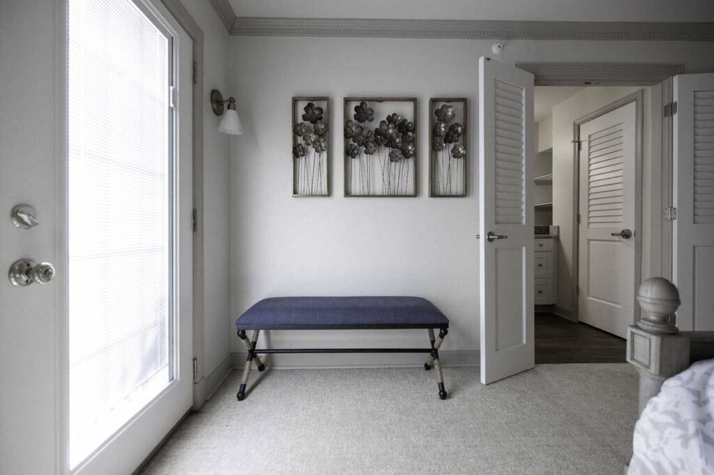 A bright room with a blue bench against a white wall, three floral art pieces above it, an open door revealing a closet, and a glass door on the left letting in natural light.