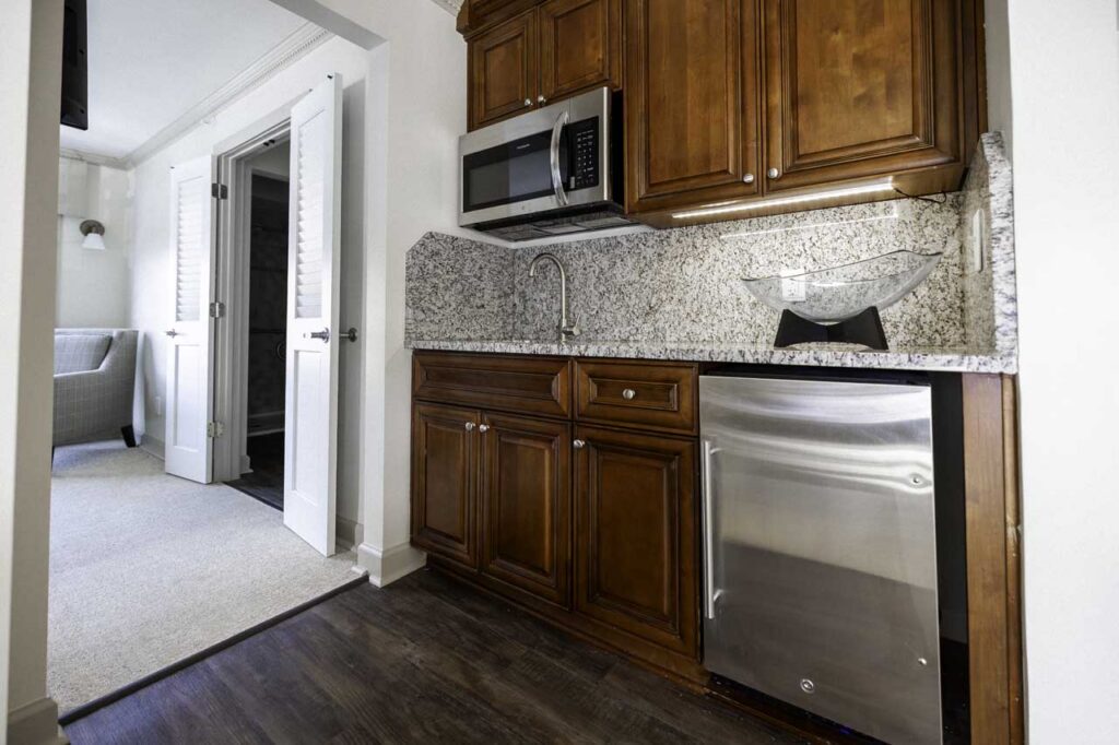 A modern kitchenette with wooden cabinets, granite countertop, stainless steel microwave and dishwasher, a glass vessel sink, and a view into an adjacent room with double doors and an armchair.