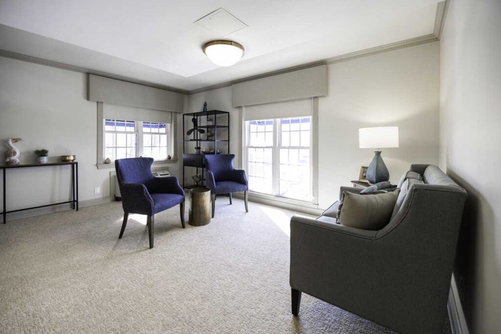 A bright, modern living room with a gray sofa, two blue armchairs, a black shelving unit, and a side table. Large windows let in natural light, and a lamp sits on a side table next to the sofa.