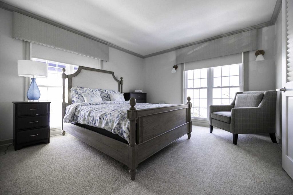 A bright bedroom with a large wooden bed, patterned bedding, a dark nightstand with a blue glass lamp, an upholstered armchair, and two large windows letting in natural light.
