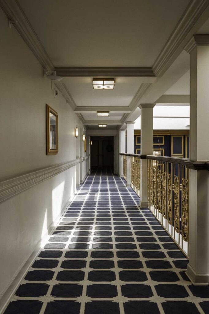 A well-lit hallway with patterned blue and white carpet, gold railing on the right, white walls, and framed artwork on the left, leading to a darker doorway at the far end.