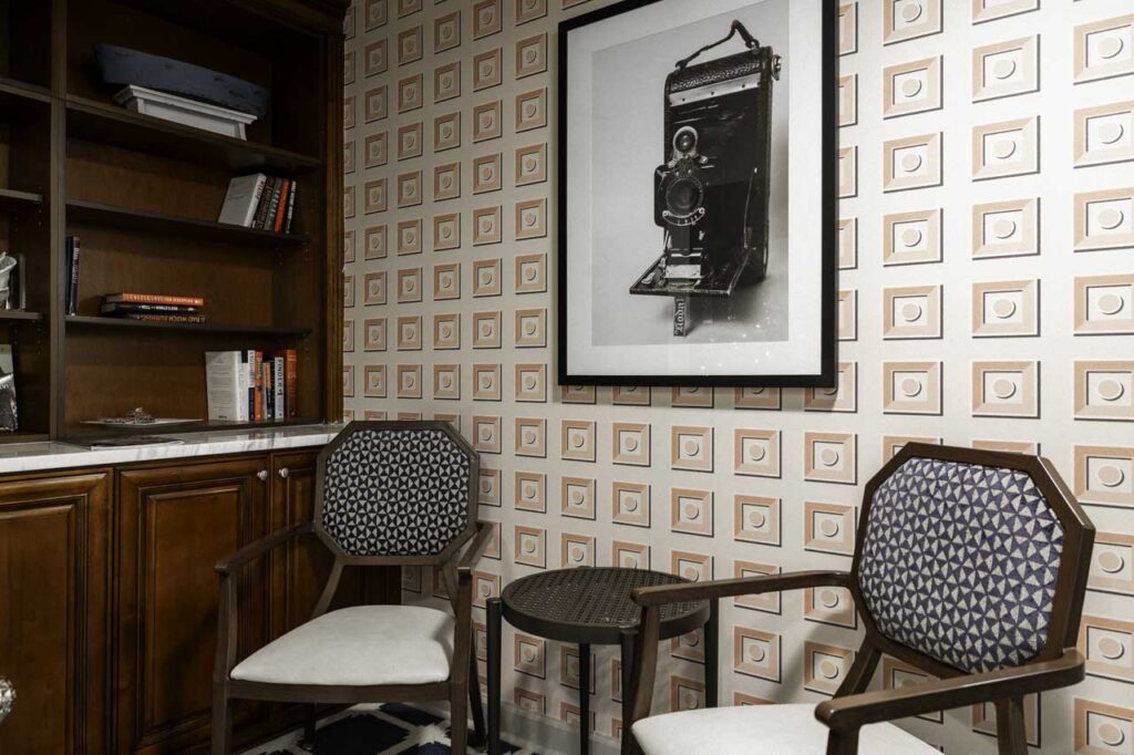 Two wooden chairs with patterned cushions sit beside a small round table, in front of wallpaper with a geometric design. A framed vintage camera photo hangs on the wall, and a bookshelf is partially visible on the left.