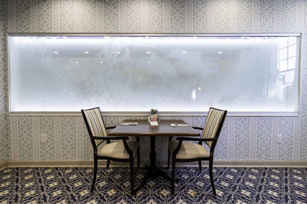 A small dining table with two chairs sits against a patterned wall with a large frosted glass panel above it. The table is set for two, with a small potted plant centerpiece. The carpet features a blue and white design.