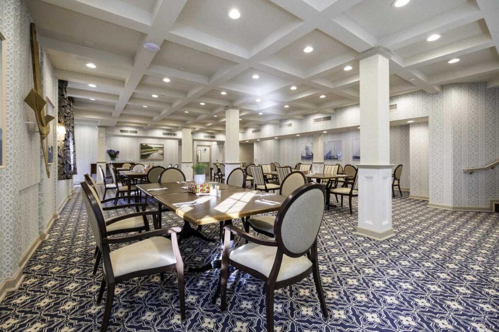 Bright, spacious dining room with patterned carpet, several wooden tables and cushioned chairs. The walls are decorated with framed artwork, and recessed lighting illuminates the room. No people are present.