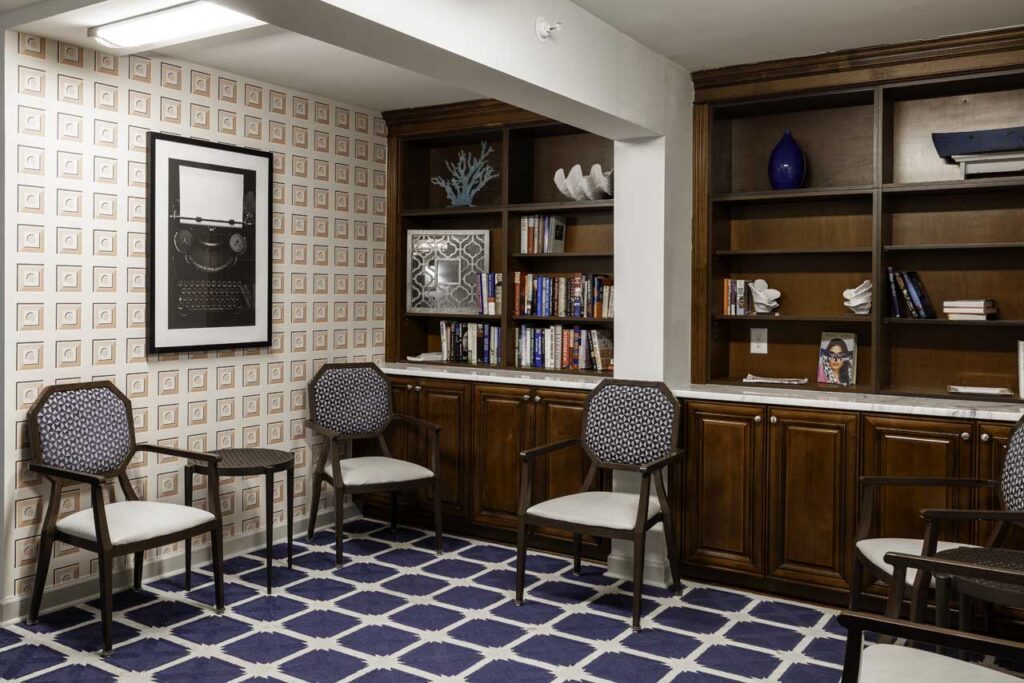 A cozy reading room with four cushioned chairs, wooden bookshelves filled with books and decorative items, a patterned carpet, and framed art on a wallpapered wall.