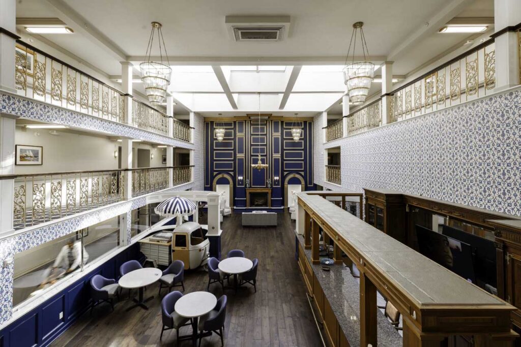 Spacious, elegant room with ornate blue and gold walls, high ceilings, patterned wallpaper, large windows, round tables with chairs, a wooden bar, and decorative chandeliers. A person walks along the upper balcony.