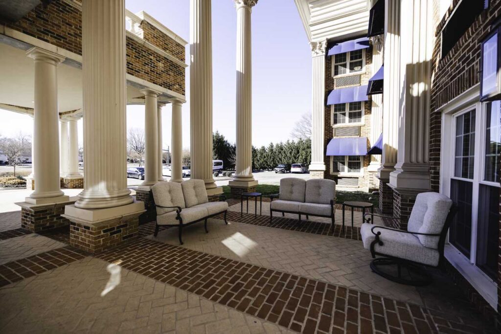 Outdoor seating area with cushioned chairs and benches arranged on a brick patio, surrounded by tall white columns and brick buildings, with parked cars and trees visible in the background.