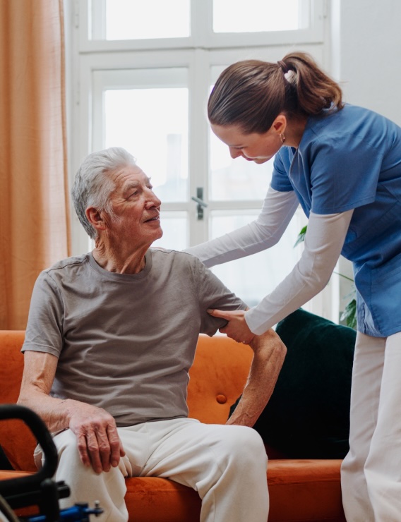 A nurse in blue scrubs helps an elderly man sitting on an orange sofa, gently holding his arm. They are indoors, and the man looks up at the nurse with a smile.