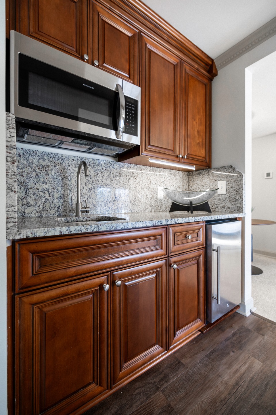 A kitchen features dark wooden cabinets, a granite countertop and backsplash, a built-in microwave, a sink with a modern faucet, and a small stainless steel refrigerator on a dark wood floor.